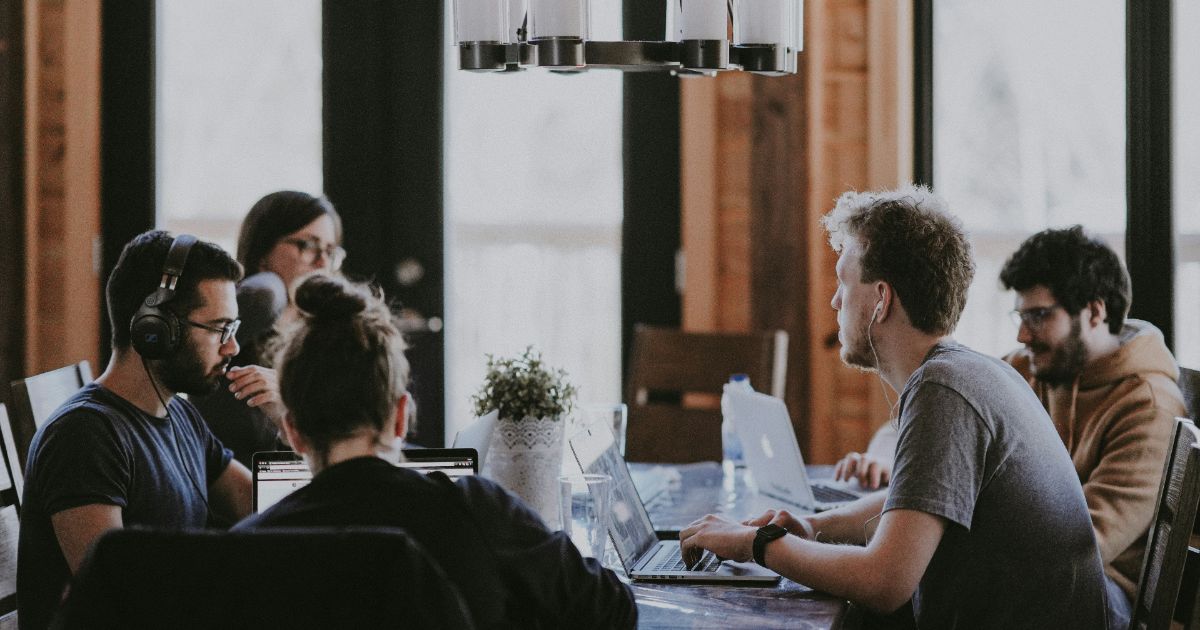 A group of five people working on laptops around a wooden table in a bright room.