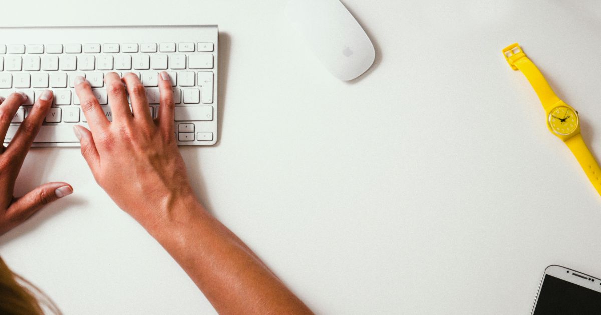 Hands typing on a white keyboard with a white mouse, a yellow wristwatch, and a smartphone on a white desk representing UX research to UX strategy.