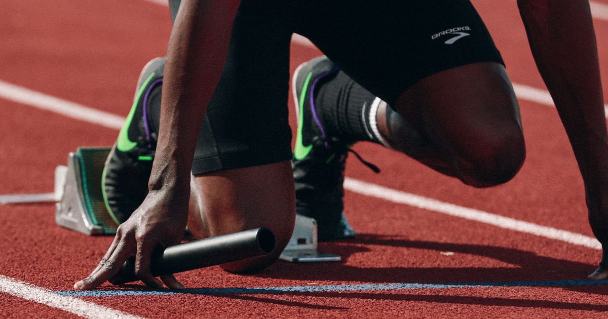 Athlete in starting position on a track, holding a baton, wearing black running gear and sneakers.