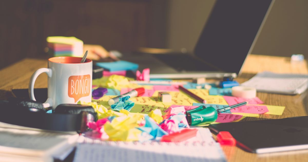 Cluttered desk with scattered colorful papers, sticky notes, pens, a mug labeled "BONGÜ," and an open laptop.