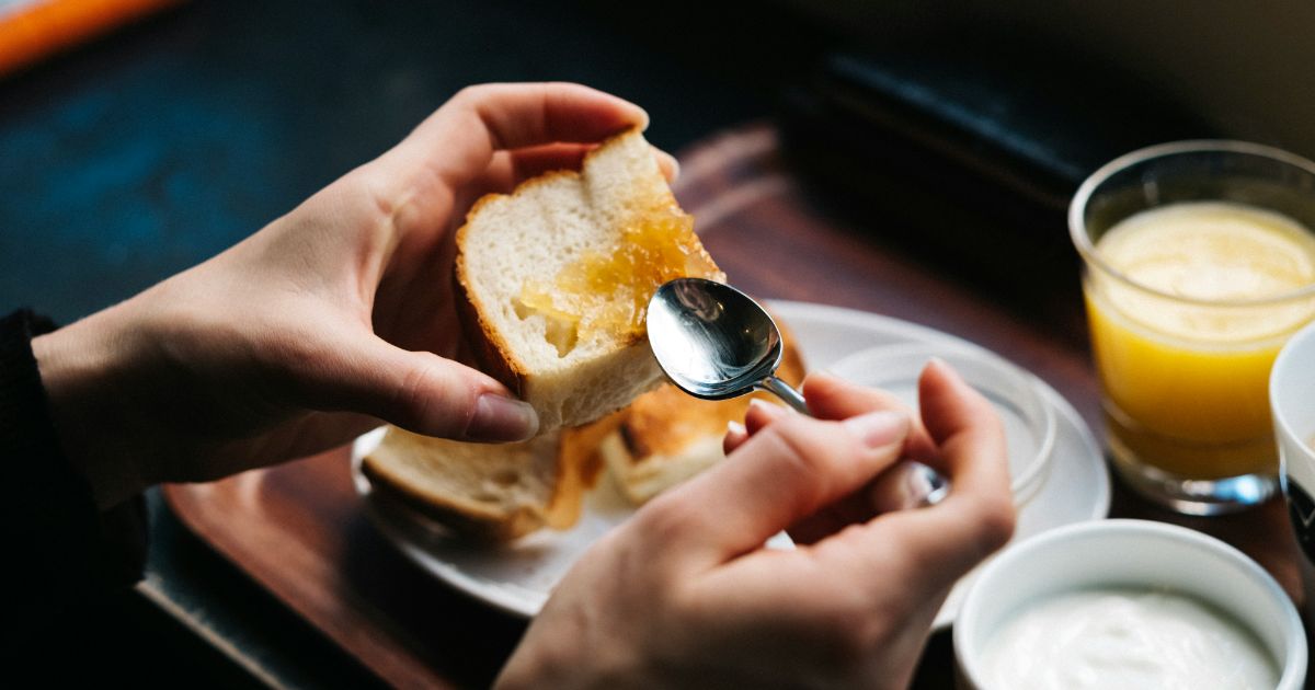 Hands spreading orange marmalade on white bread with a spoon, next to juice and yogurt.