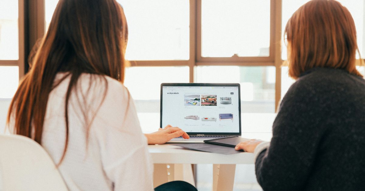 Two people sitting at a table looking at a laptop displaying a furniture website.