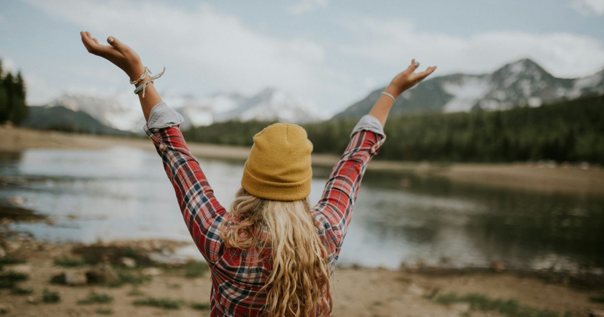Person in a yellow beanie and red plaid shirt with arms raised, standing by a lake with mountains in the background.