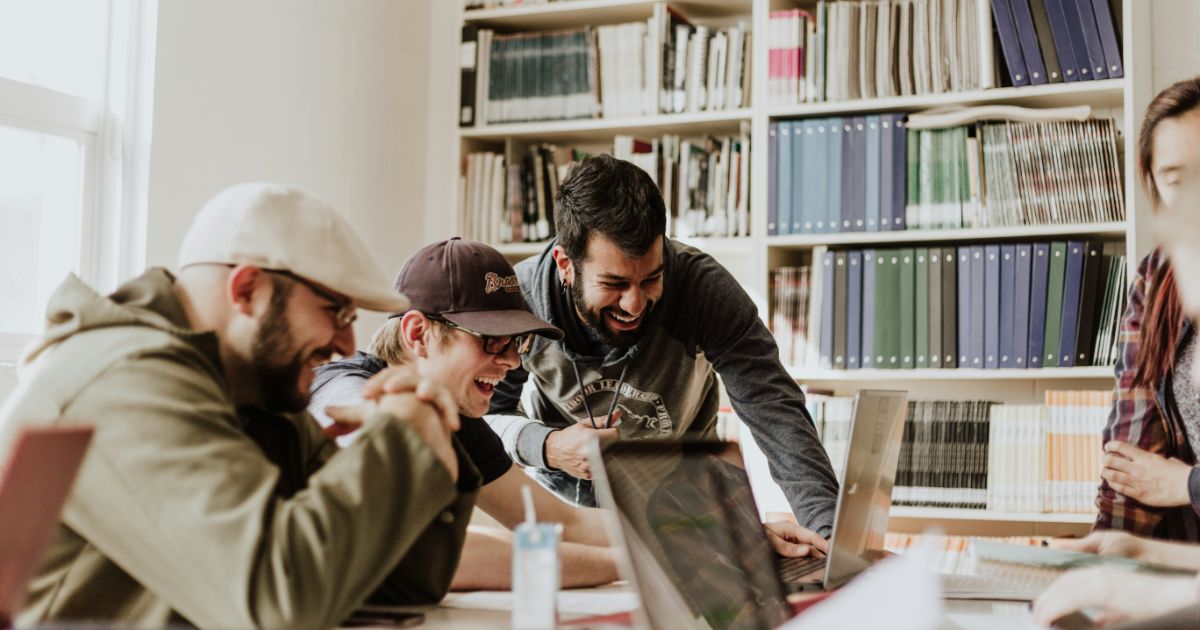 Group of four people laughing around a laptop in a library setting.