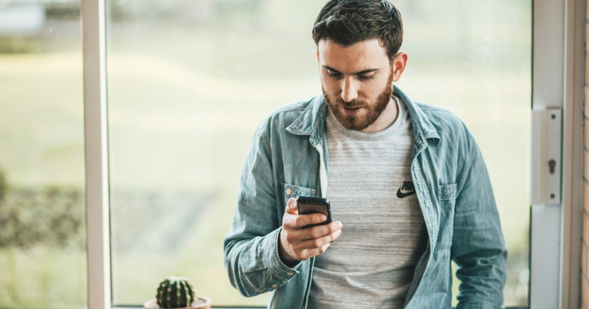 Man looking at a smartphone by a window with a small cactus on the windowsill.