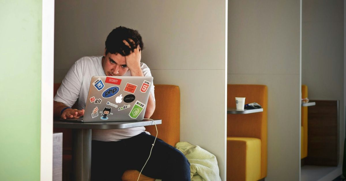 A frustrated designer sits in a modern workspace, staring at his laptop covered in coding and tech stickers like PHP, Git, and HTML5. This image reflects the challenges faced when trying to implement user-centered design principles, especially when creating intuitive, user-friendly interfaces. It highlights the importance of understanding UCD basics, involving users early, and iterating designs to avoid design pitfalls that lead to user frustration.