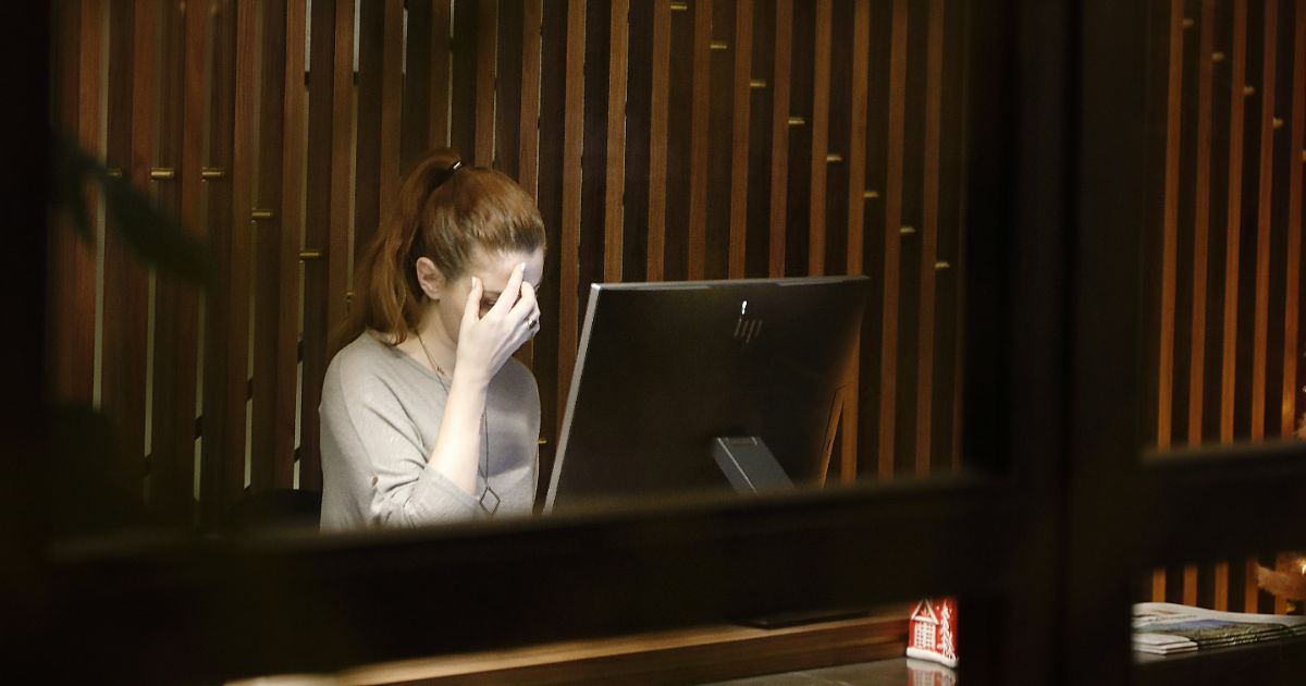 Woman at a desk, viewed through a window, holds her head in front of a computer monitor, with a wooden partition behind her.