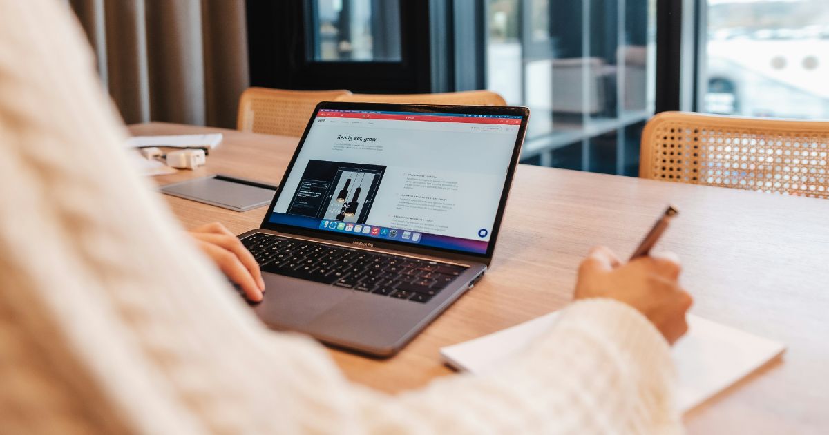 Person using a laptop at a wooden table with a notepad and pen.