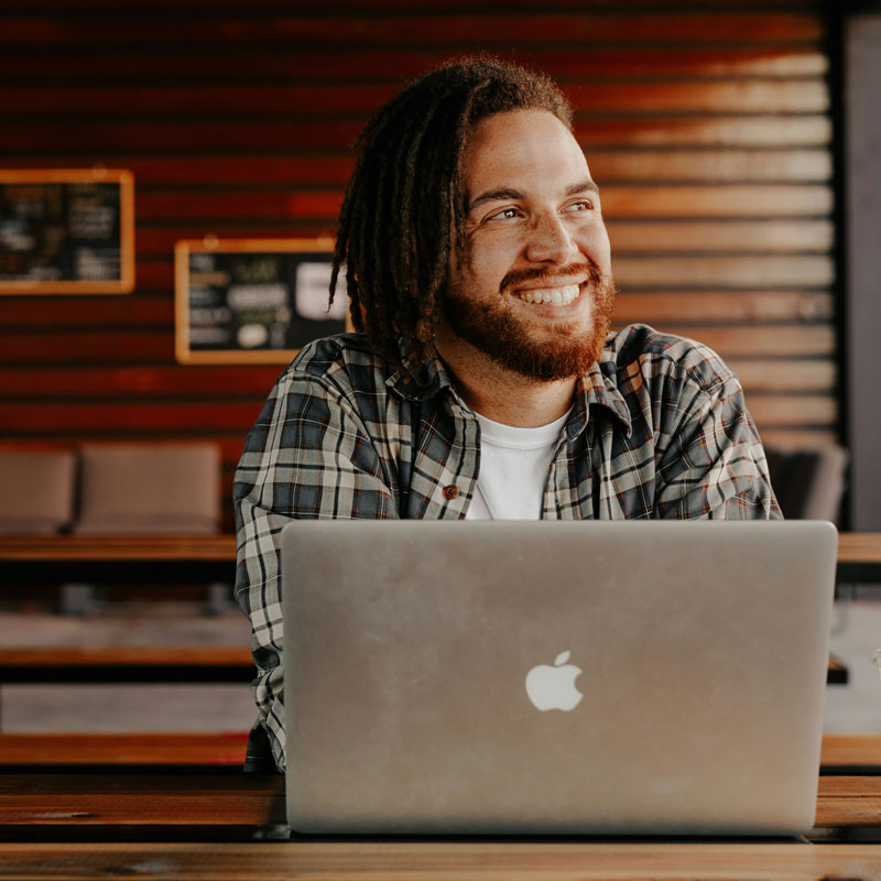 brooke-cagle-Rat0xIB5X1c-unsplash A man with dreadlocks sits at a table, focused on his Apple laptop, creating a relaxed and productive atmosphere.