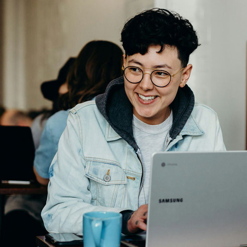 brooke-cagle-_6cz9KUvnxM-unsplash A smiling woman in glasses and a denim jacket is happily using her laptop.