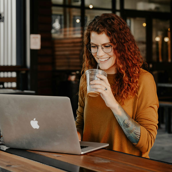brooke-cagle-cb4Dv50LN1Y-unsplash A woman with red hair and glasses is seated at a table, focused on her laptop.