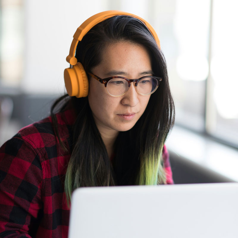 christina-wocintechchat-com-AHfRjkk8QcE-unsplash A woman in a plaid shirt wears headphones while focused on her laptop, immersed in her work.