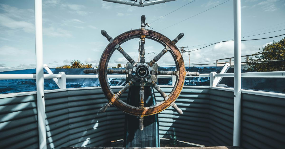 A close-up of a boat steering wheel, showcasing its wooden texture and nautical design against a blurred boat background.