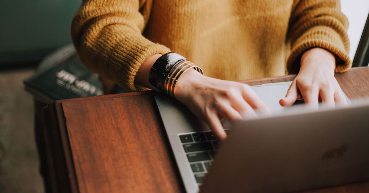 Person wearing a mustard sweater typing on a silver laptop with books nearby on a wooden table.