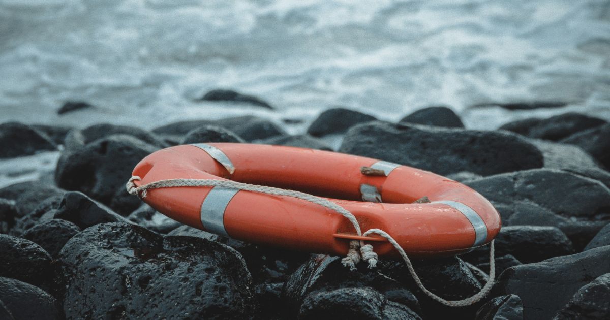 An orange life preserver rests on a rocky beach, contrasting with the gray stones and the blue ocean in the background symbolizing universal design in disaster response.