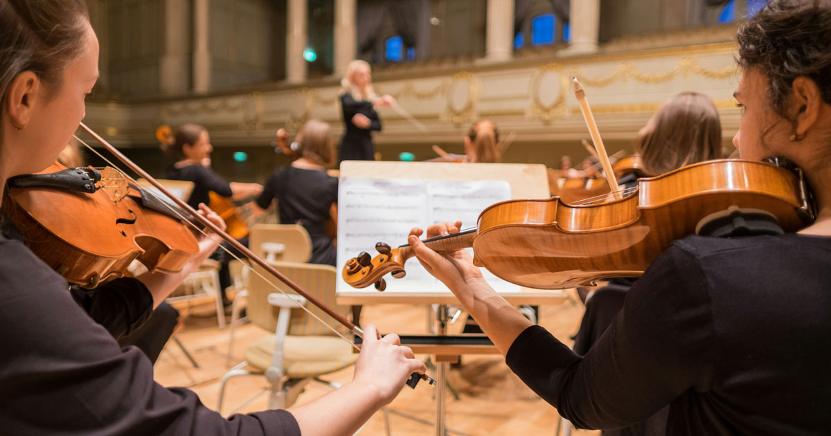 A group of young women performing together, playing violins in an orchestra setting, showcasing their musical talent and Zero-UI trends.