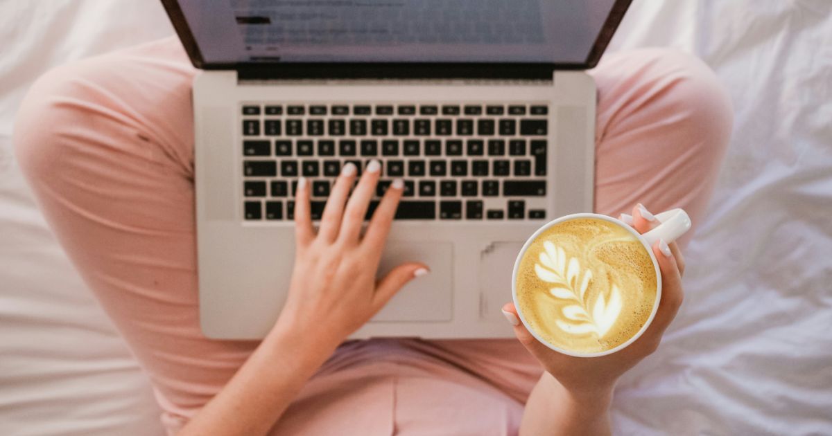 A woman sits on a bed, working on her laptop while enjoying a latte beside her representing information architecture for multi-entry journeys.