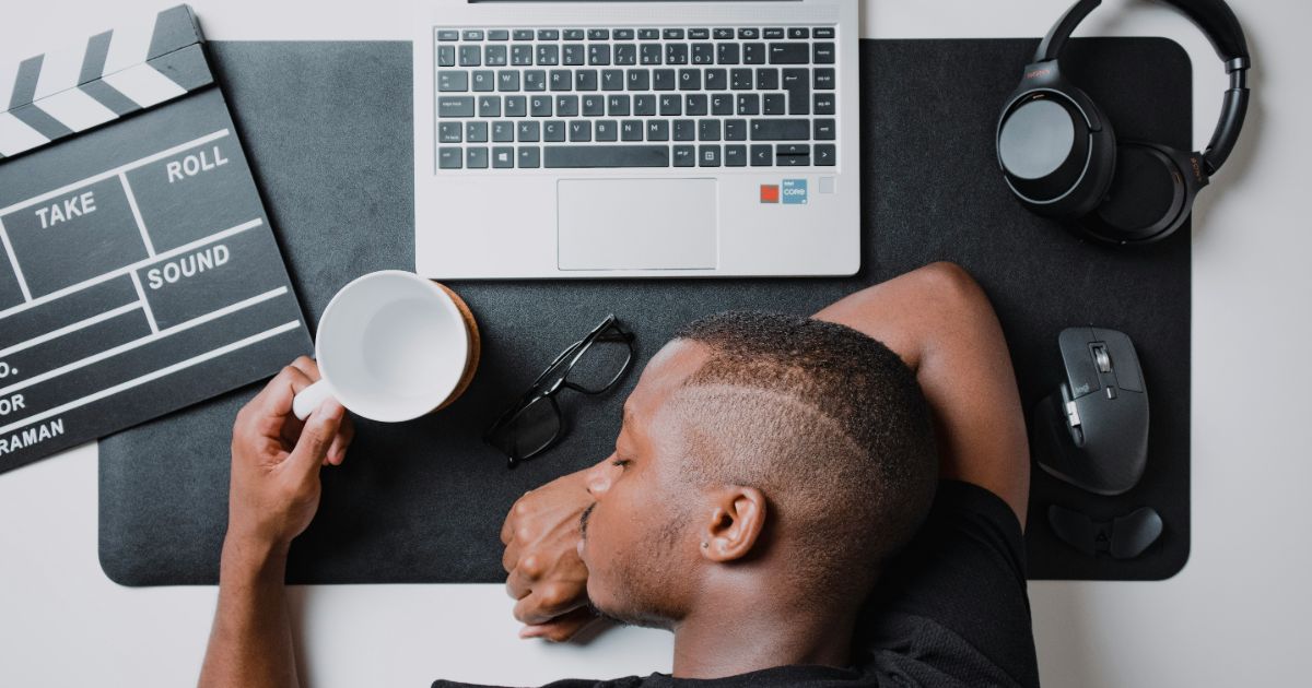 A man is sleeping with his head resting on a laptop, next to a coffee cup on a table representing designing for digital exhaustion.